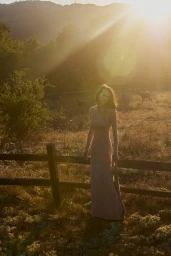 Woman in a long dress standing by a wooden fence in a meadow during sunset, captured by Lindsey Childs.