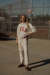 Woman in an oversized USA sweatshirt and light trousers standing on an outdoor asphalt court by a chain-link fence