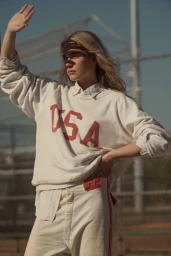 Blonde woman wearing white USA sweatshirt raising her hand to block the sun against an outdoor chain-link fence.