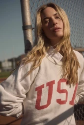 Blonde woman wearing white USA sweatshirt leaning against chain-link fence outdoors in sunlight