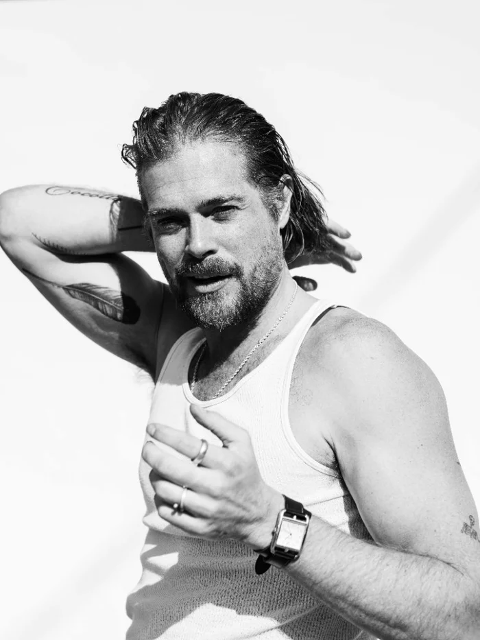 Monochrome portrait of a man with dark hair in a white tank top wearing rings and a wrist watch against a bright background