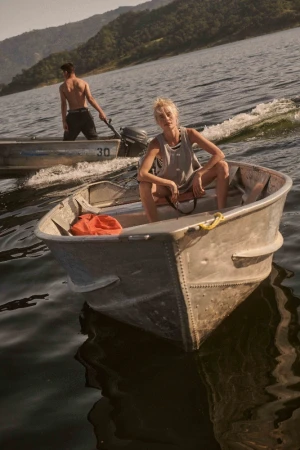 Blonde woman seated in an aluminum motorboat on a lake with a shirtless man standing in another boat behind her.