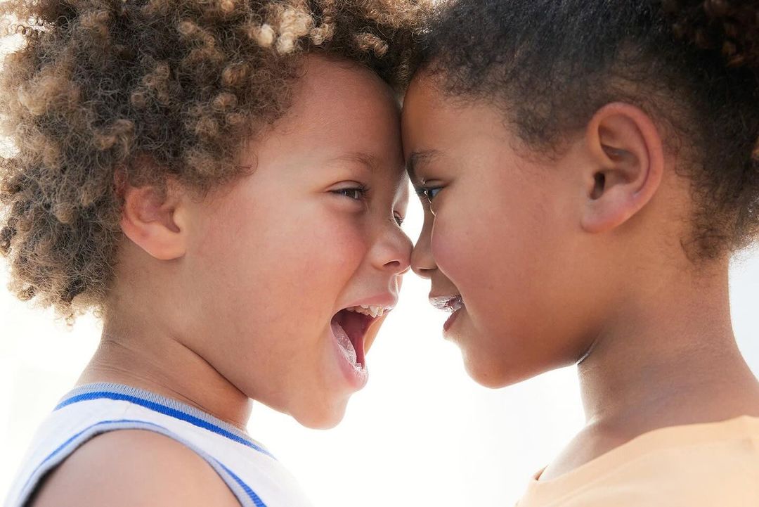 Two young children, one boy and one girl, are face to face, foreheads touching. The boy is mid-laugh with his mouth open, showing his tongue.
