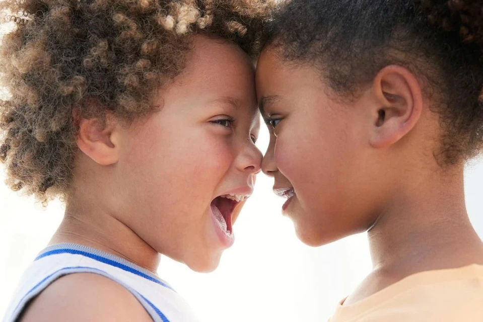 Two young children, one boy and one girl, are face to face, foreheads touching. The boy is mid-laugh with his mouth open, showing his tongue.