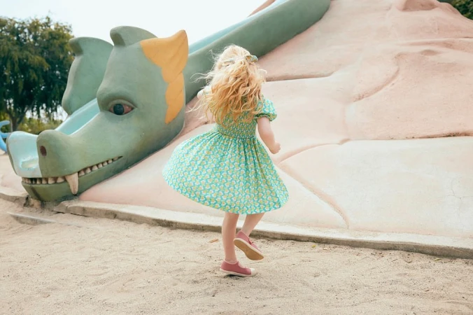 Young girl in a patterned dress twirling in front of a large, green dragon sculpture in an outdoor park on a sunny day.