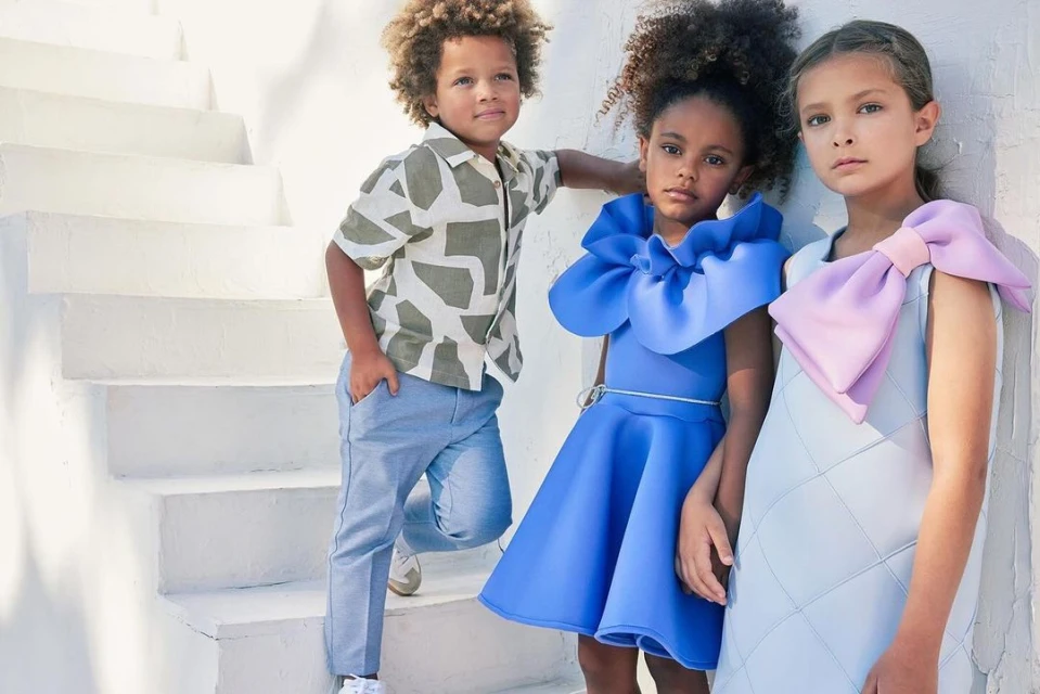 Three children pose on white steps. One boy wears a patterned shirt and blue pants, and two girls wear blue and light blue dresses with bows.