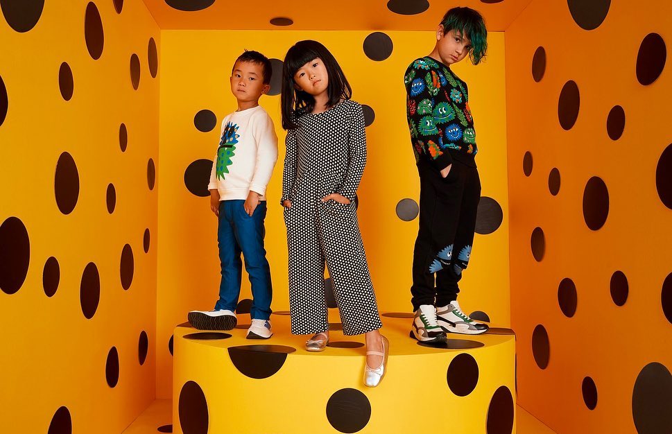 Three stylish children's fashion models pose in a bright yellow polka-dot room, showcasing a modern and playful aesthetic in a studio setting.