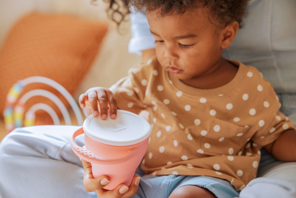 African American toddler sits on an adult's lap and plays with a pink and white lidded cup in a bright indoor setting.