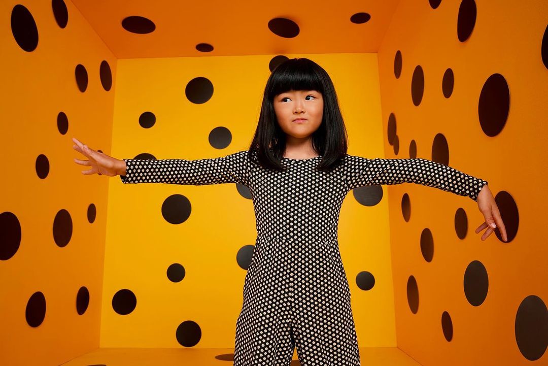 A young Asian girl in a polka-dot jumpsuit stands in a yellow room covered in black polka dots, arms outstretched. 