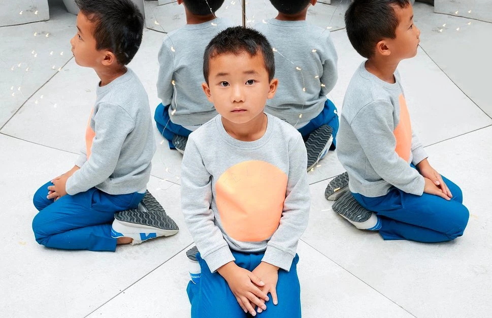 Child in a gray sweatshirt and blue pants kneels indoors with reflective surfaces showing multiple views of the same child.