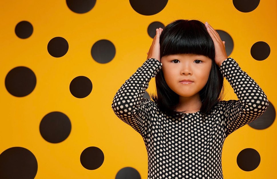 Young girl with hands on head poses in front of an orange background with black polka dots, wearing a black and white polka dot dress.