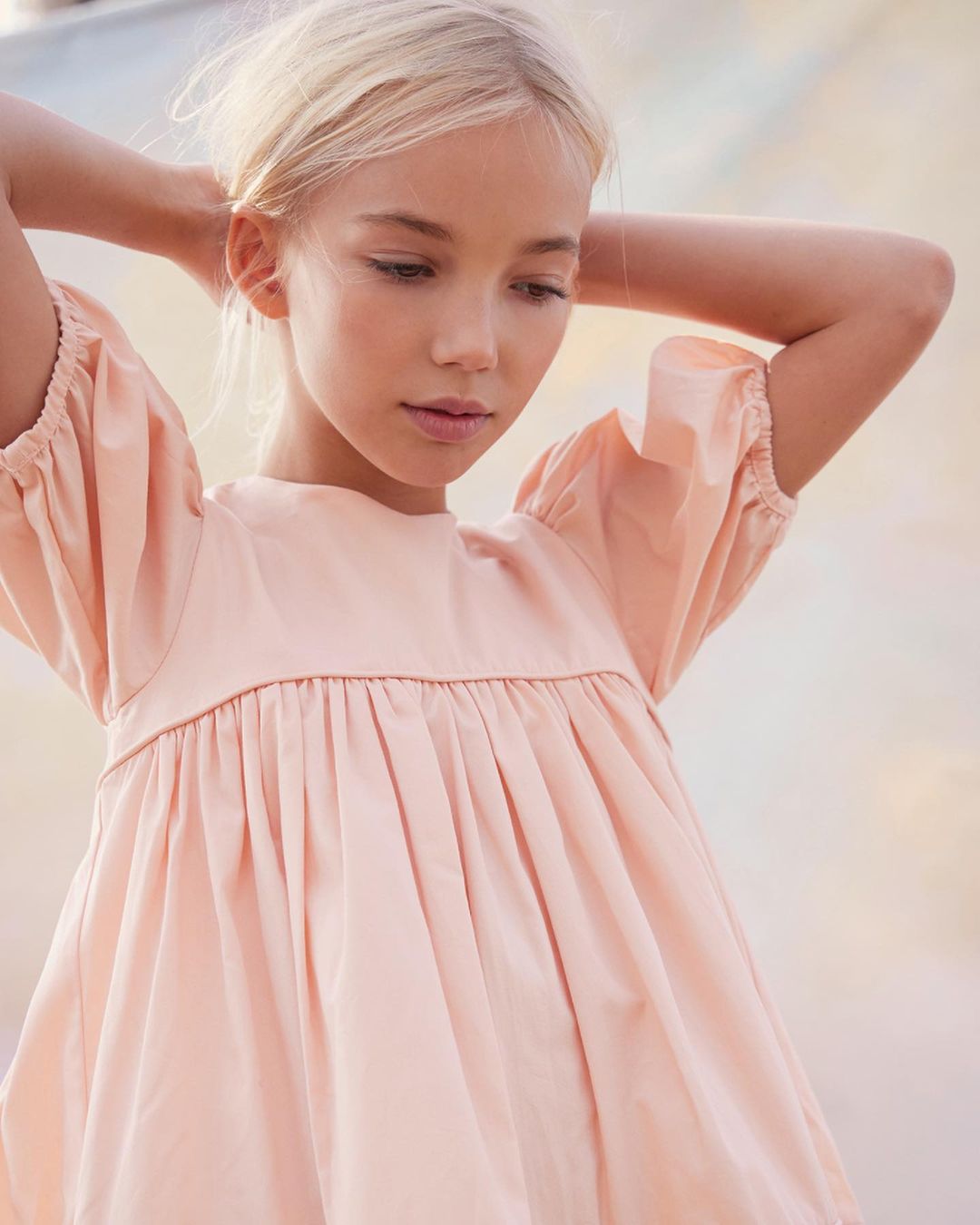 Close-up studio shot of a young blonde girl in a pink dress with her hands behind her head looking down, conveying a soft, dreamy mood.