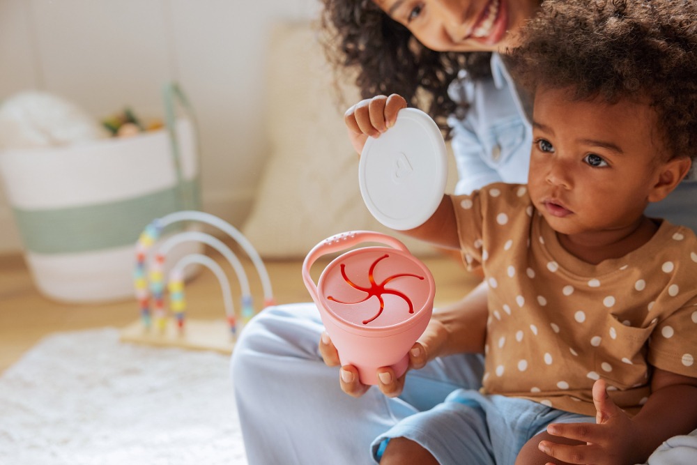 Young child sits indoors with mother, holding a pink snack container with a white lid, on a white rug with a rainbow toy in the background.