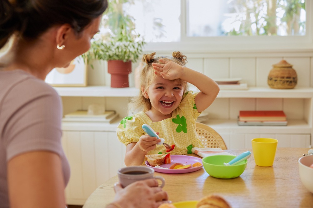 Smiling toddler with hand on forehead sits at a wooden table with a woman and colorful dishes, enjoying food and a beverage indoors.