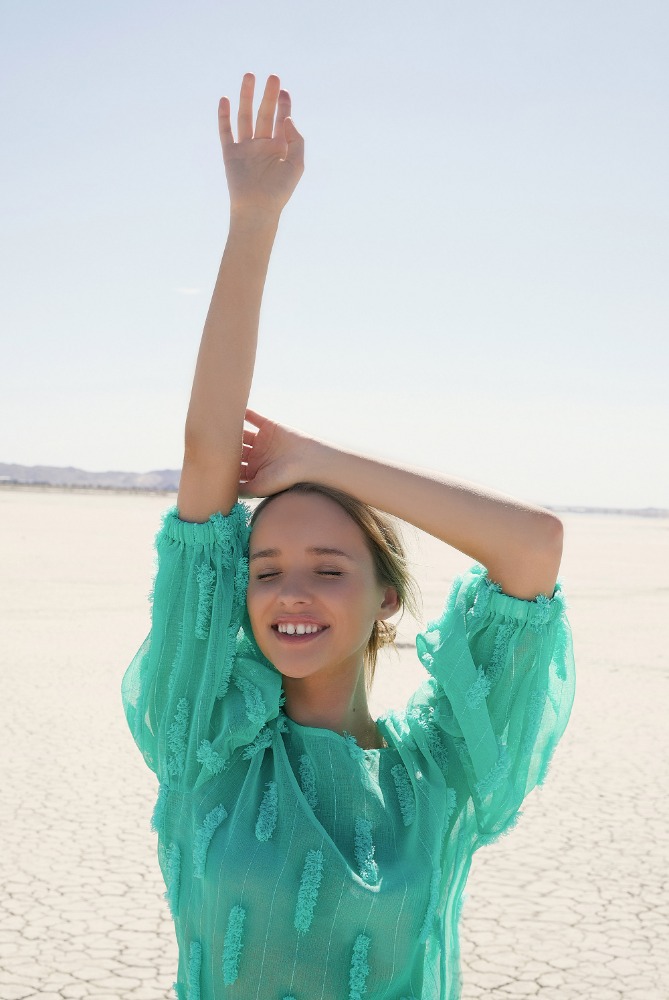Woman in turquoise blouse with arms raised outdoors on a dry, cracked lakebed under a clear sky, expressing happiness and freedom.