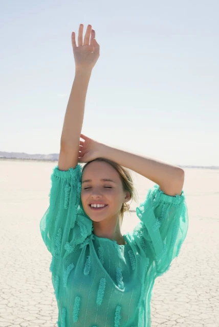 Woman in turquoise blouse with arms raised outdoors on a dry, cracked lakebed under a clear sky, expressing happiness and freedom.