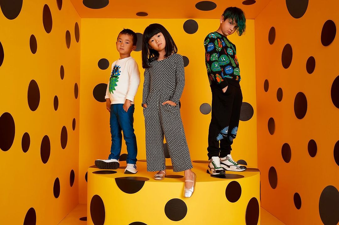 Three kids in modern clothing pose on a polka-dot set, one stands on a cylinder in an orange room with black dots, fashion.