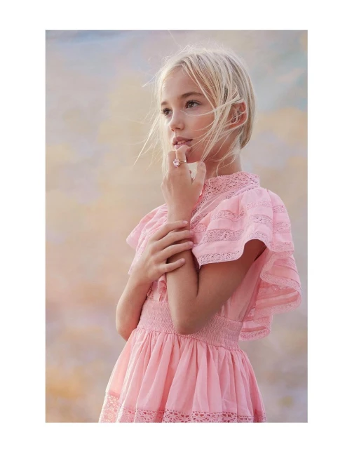 Blond girl in pink lace dress looks away against a blurred sky background, posing with her hands near her face, soft focus, portrait.