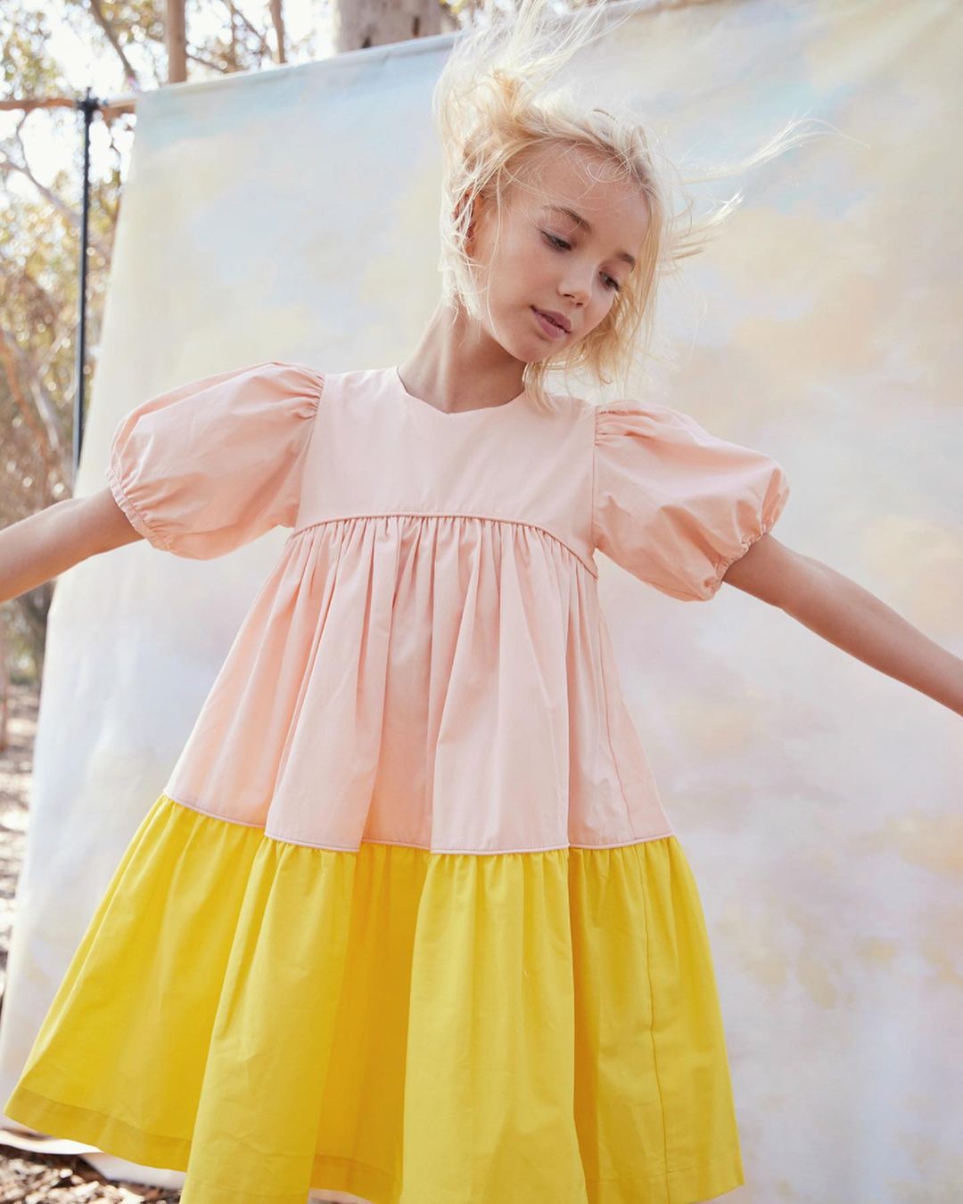 A young girl with blonde hair wears a pink and yellow dress with puff sleeves. She is outdoors in a blurred natural environment.