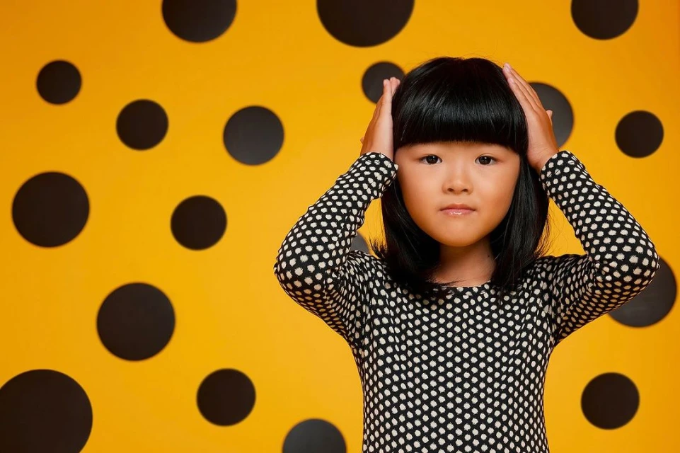 Young Asian girl with hands on her head poses in front of a patterned yellow and black background wearing a polka dot top.