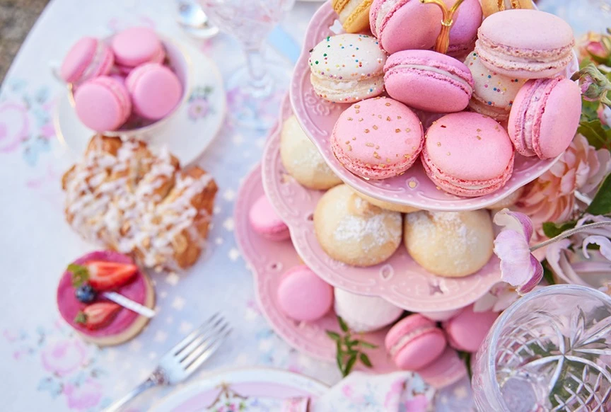 Overhead shot of a pink dessert tower topped with macarons, surrounded by pastries, flowers, and vintage tableware, capturing a pastel aesthetic.