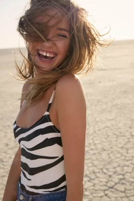 A woman laughs in a zebra-striped bodysuit on a cracked desert landscape. Golden hour lighting highlights her hair and skin.
