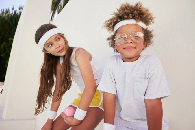 Two children wearing retro sports apparel and white headbands posing outdoors against a plain wall.