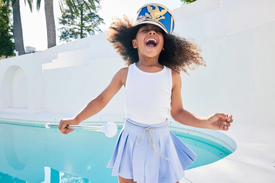 Young girl in a majorette costume jumps joyfully by a turquoise swimming pool outdoors on a sunny day, wearing a blue drum major hat.