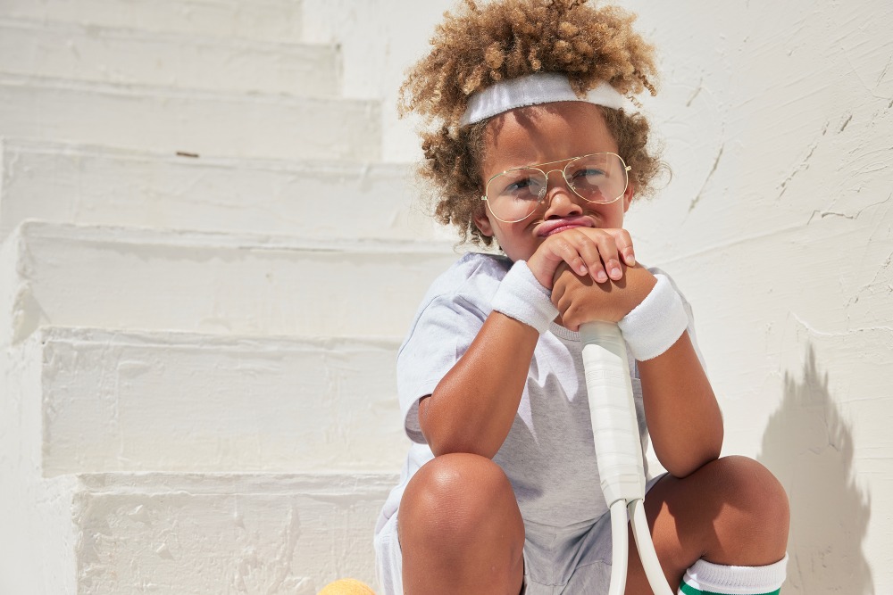A young child with curly hair and glasses sits on white stairs holding a tennis racket, dressed in sportswear with a pouty facial expression.
