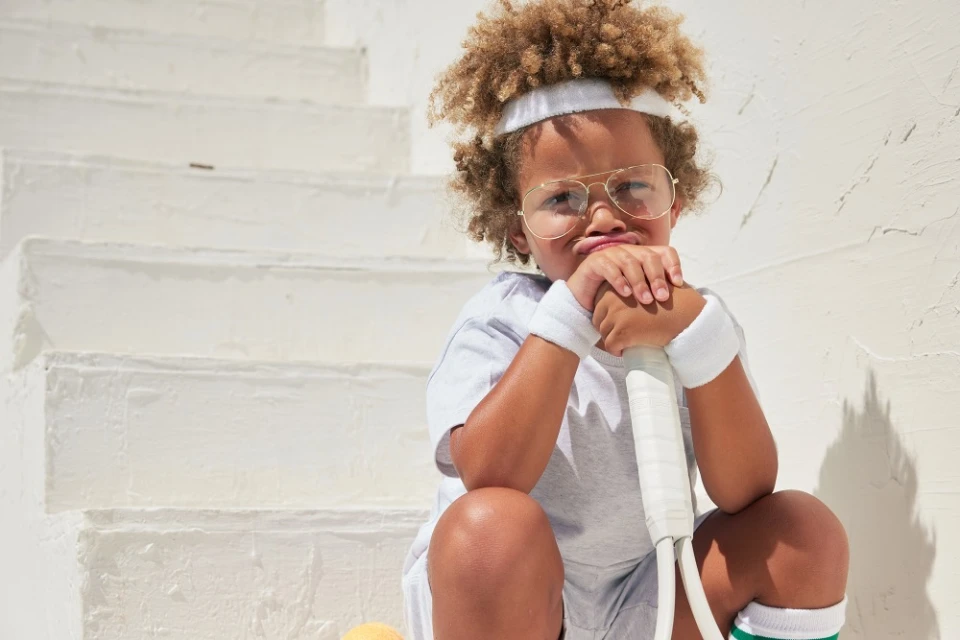A young child with curly hair and glasses sits on white stairs holding a tennis racket, dressed in sportswear with a pouty facial expression.