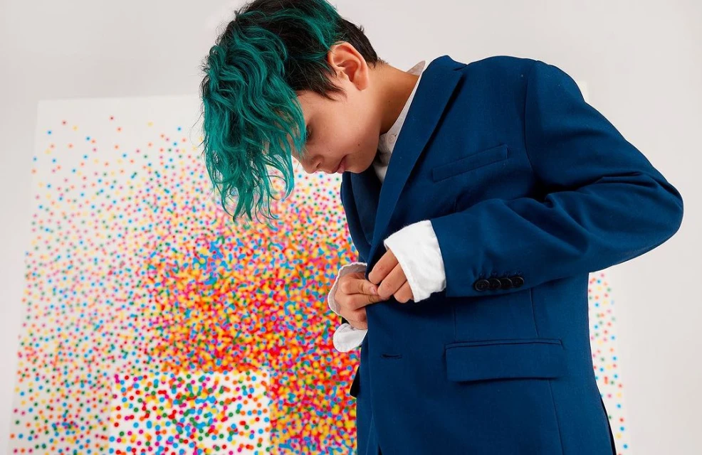 Young adult with teal hair wearing a blue blazer fastens the button, standing in front of a colorful abstract dot pattern on a white wall.
