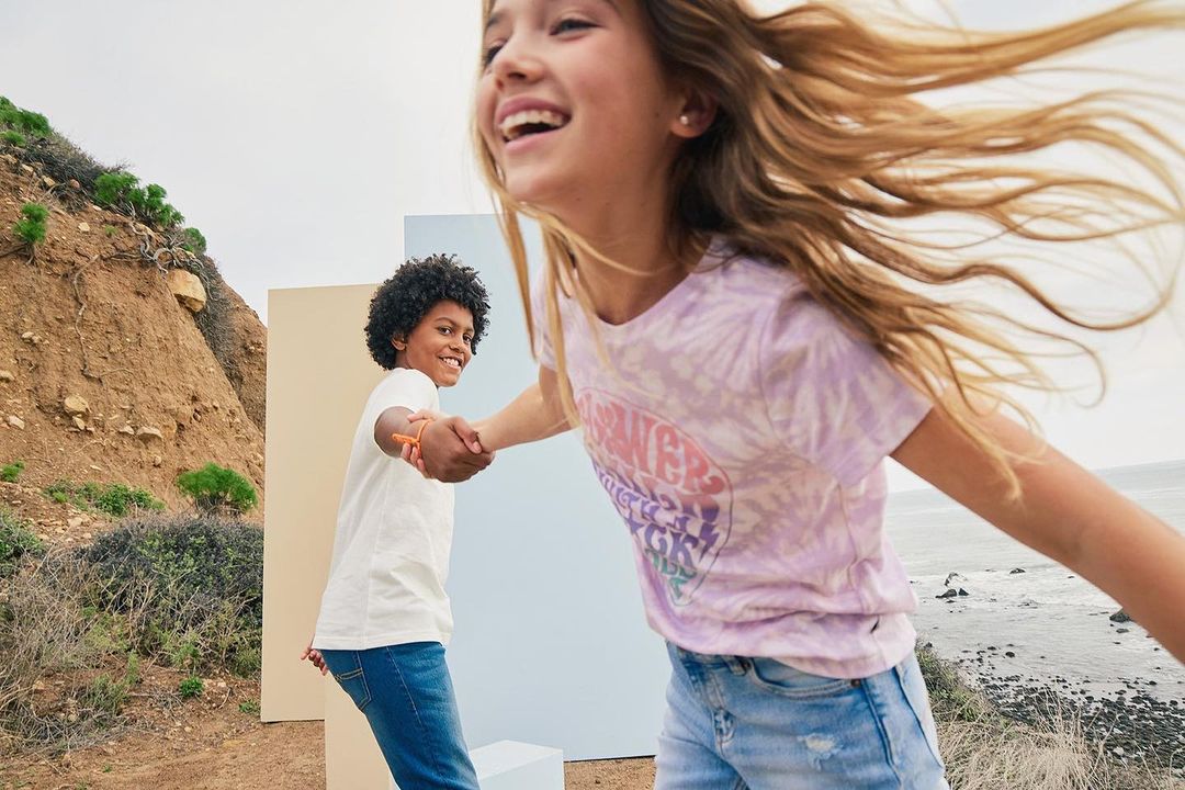 Two young people holding hands on a ridge path with ocean view. The girl wears a purple shirt and the boy a white tee with blue jeans.