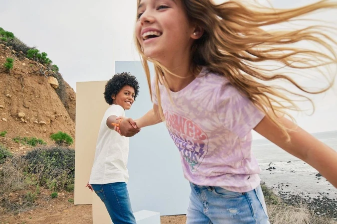 Two young people holding hands on a ridge path with ocean view. The girl wears a purple shirt and the boy a white tee with blue jeans.