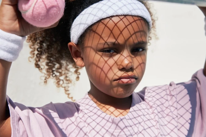 Close-up of a girl holding a tennis ball, wearing a white headband and wristband, with a patterned shadow cast on her face.