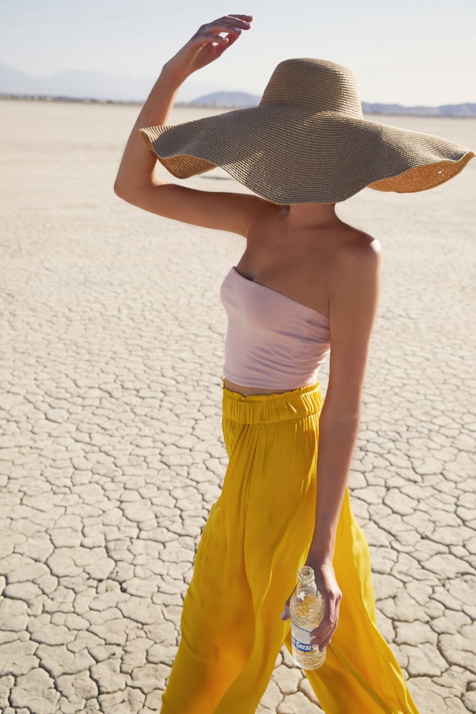 Woman in pink top and yellow pants walks across dry cracked earth wearing a large straw sun hat and holding a bottle of water.