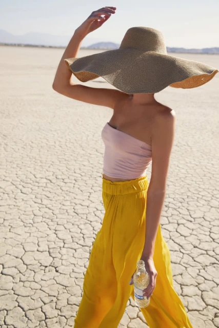 Woman in pink top and yellow pants walks across dry cracked earth wearing a large straw sun hat and holding a bottle of water.