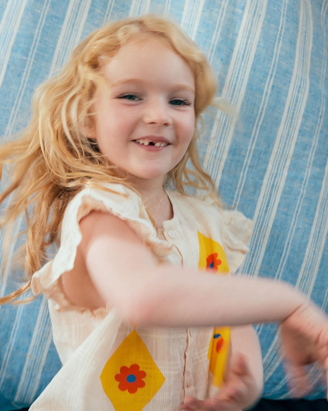 Blonde girl smiling and twirling in front of a striped blue and white background, wearing a cream-colored dress with floral diamonds.