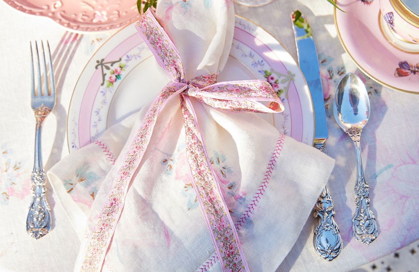 Overhead shot of ornate silverware, a floral plate, and a linen napkin with pink floral ribbon, arranged on a table setting.
