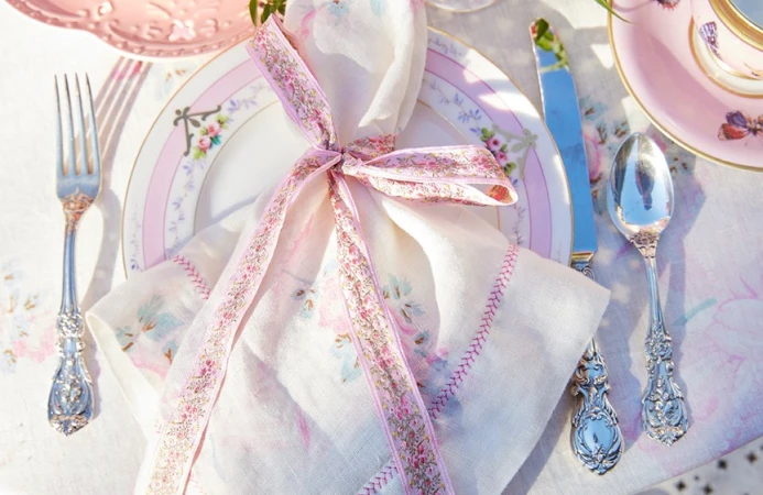 Overhead shot of ornate silverware, a floral plate, and a linen napkin with pink floral ribbon, arranged on a table setting.