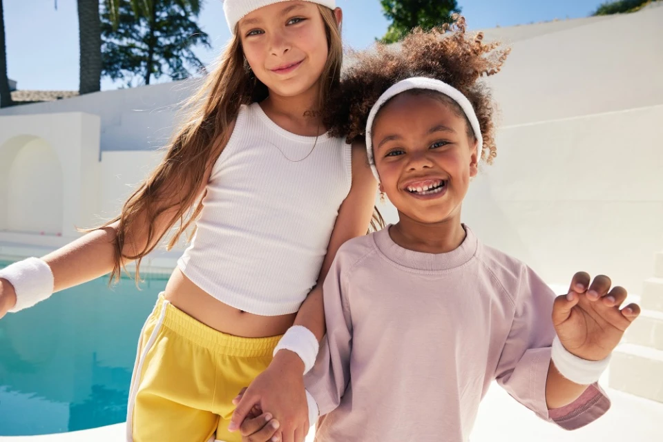 Two young girls smiling in yellow shorts and a light purple shirt, wearing matching headbands and wristbands, posing near a pool.