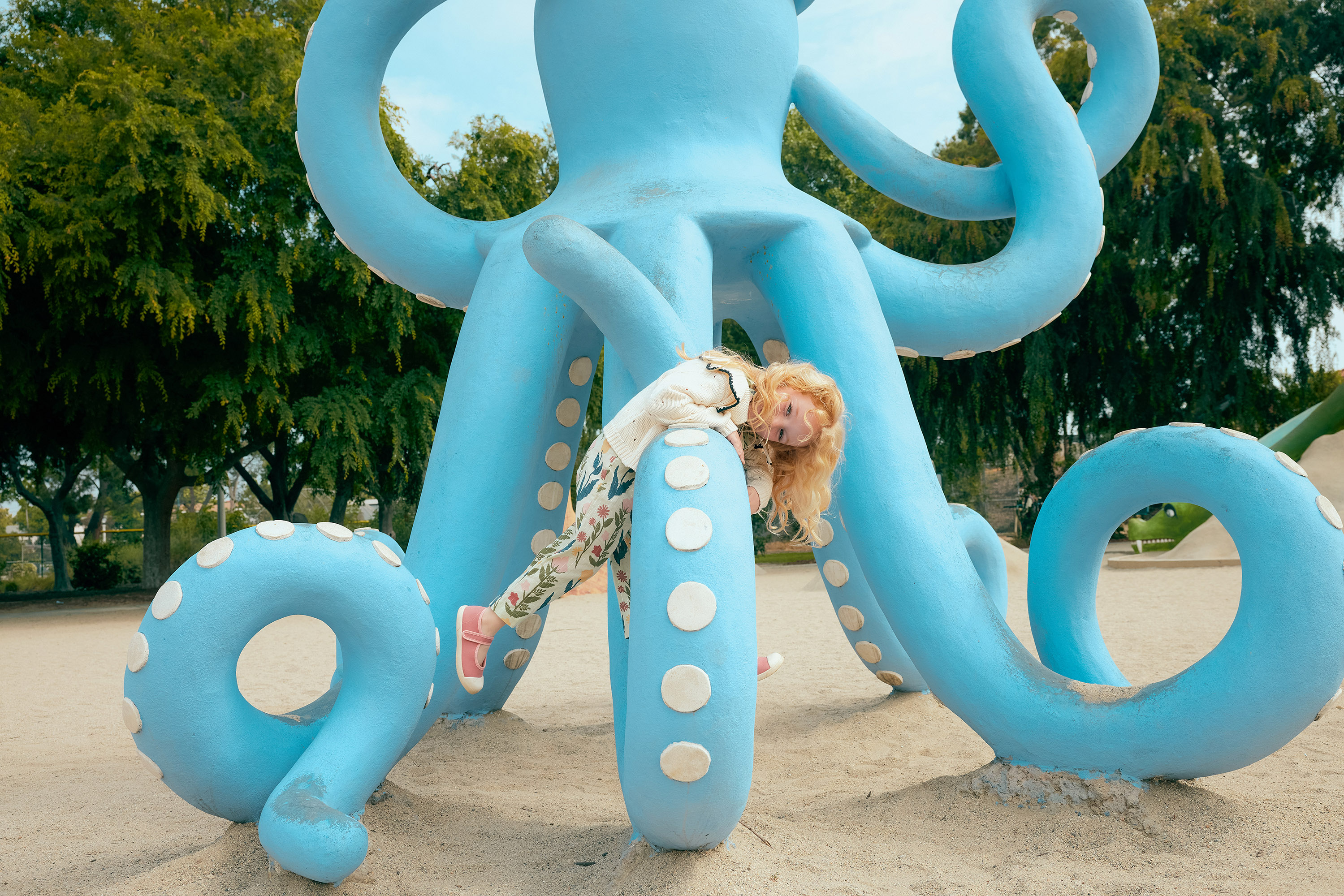 A young girl with blonde hair climbs on a large, light-blue octopus sculpture in an outdoor playground, surrounded by trees and sand.