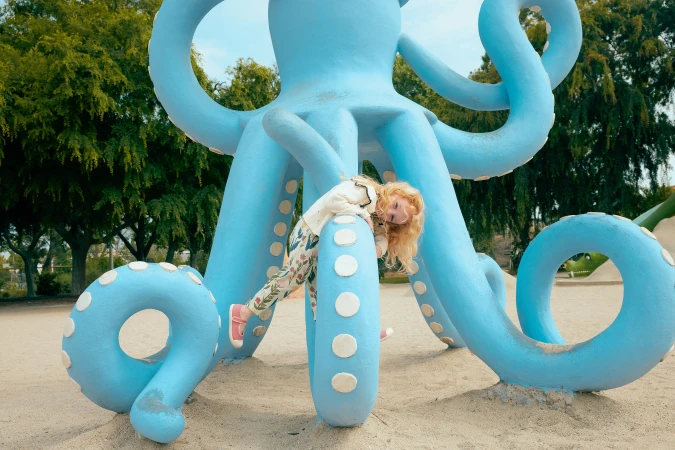 A young girl with blonde hair climbs on a large, light-blue octopus sculpture in an outdoor playground, surrounded by trees and sand.