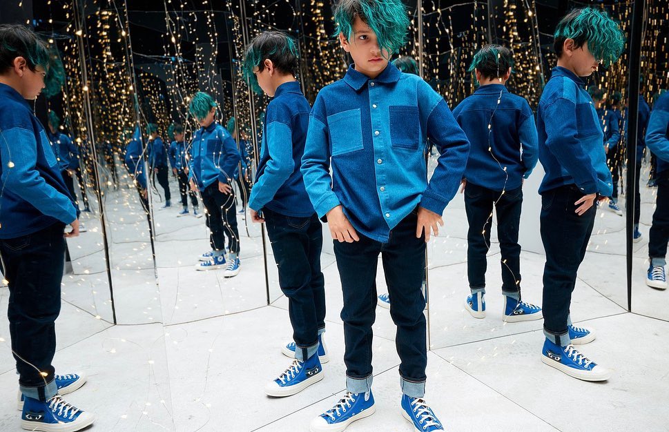 Young boy with teal hair poses in a denim shirt, black pants, and blue sneakers, surrounded by mirrors and string lights.