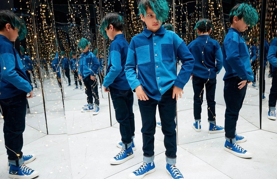 Young boy with teal hair poses in a denim shirt, black pants, and blue sneakers, surrounded by mirrors and string lights.