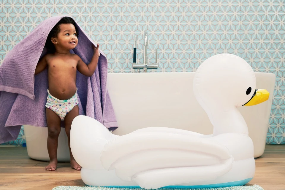 Toddler stands in a bathroom with a purple towel over their head, an inflatable swan, and patterned blue-and-white tiles.