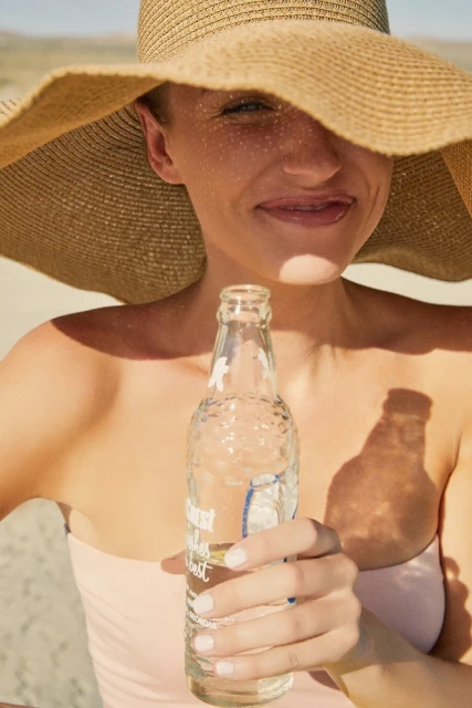 Woman in sun hat holds a glass bottle filled with clear liquid, likely water, on a sunny day at the beach.