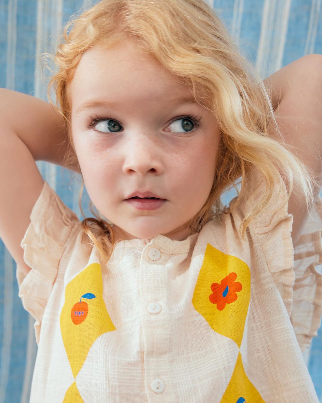 Portrait of a blonde toddler girl looking up to the left, wearing a patterned top with yellow and orange diamond shapes indoors.