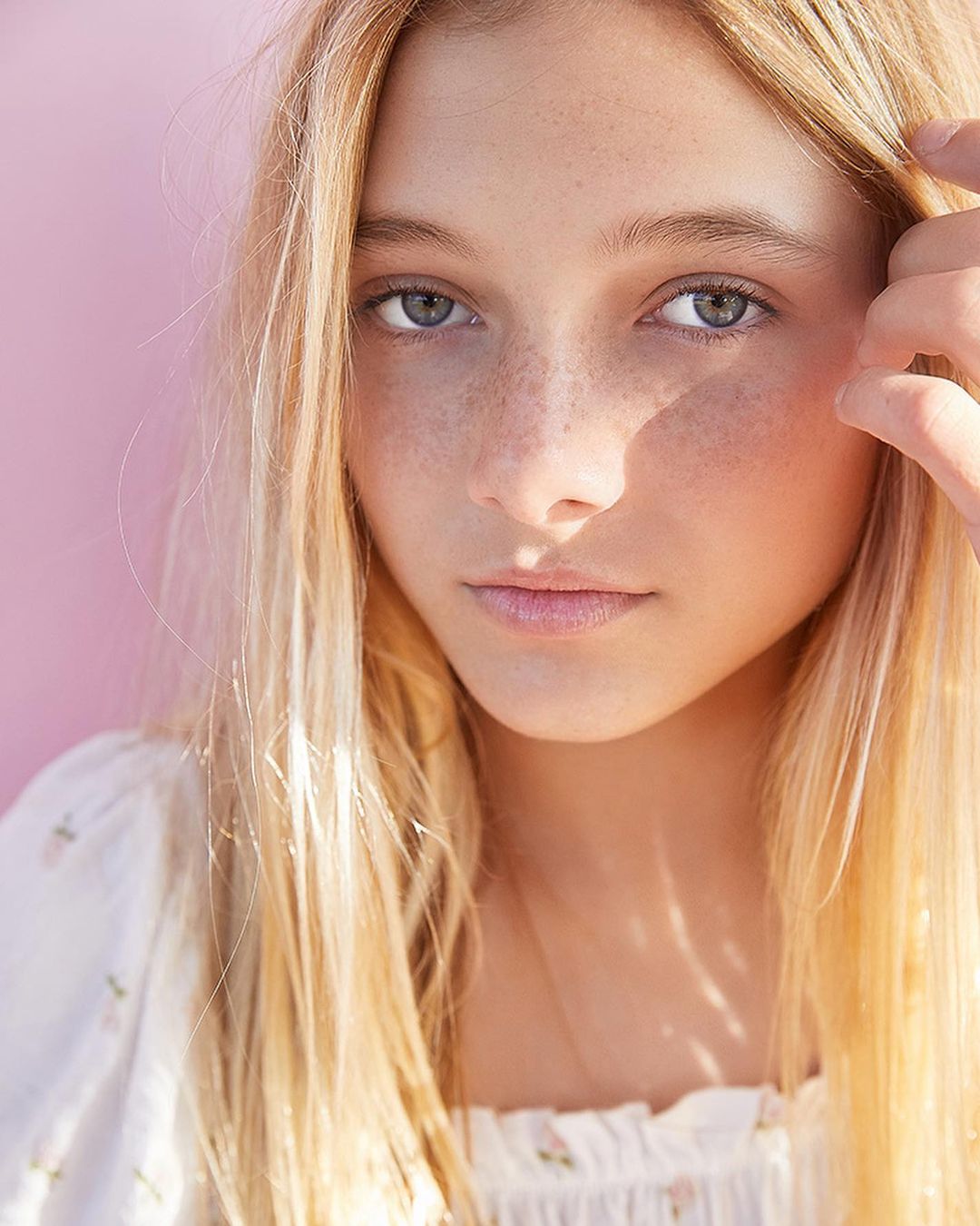Portrait of a young blonde girl with freckles, posing in front of a pink wall, wearing a white floral print top in natural light.