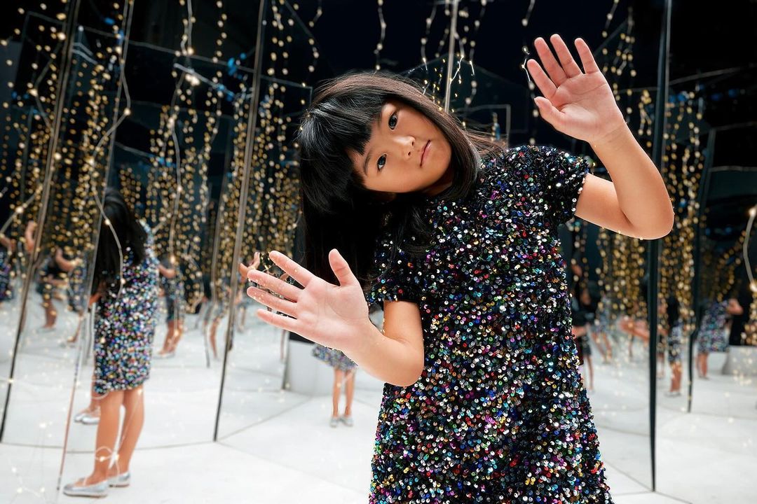 Young girl in a sequined dress poses in a room with mirrored walls reflecting fairy lights, creating a dazzling and immersive atmosphere.
