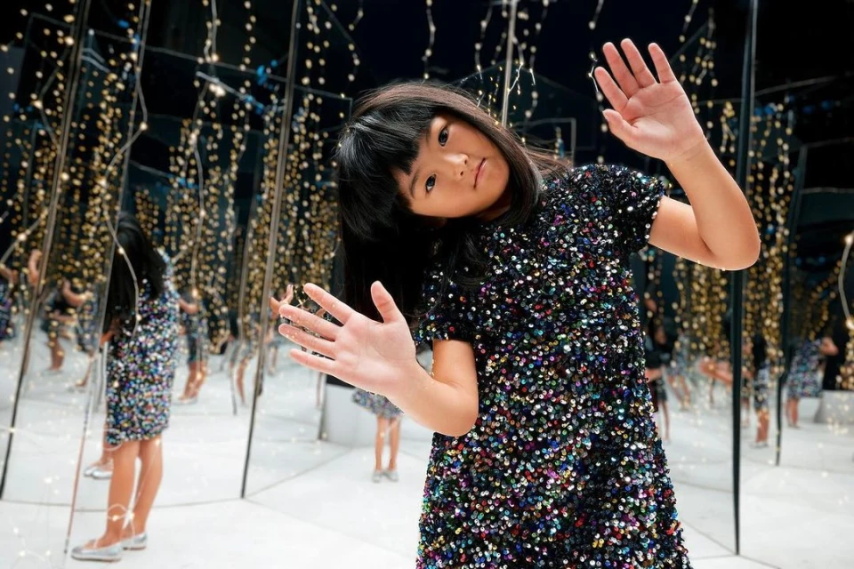 Young girl in a sequined dress poses in a room with mirrored walls reflecting fairy lights, creating a dazzling and immersive atmosphere.